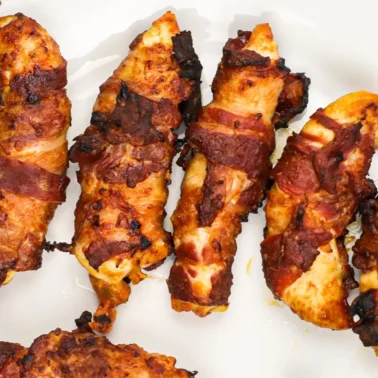 Five crispy, bacon-wrapped chicken tenders on a white plate with a patterned rim.