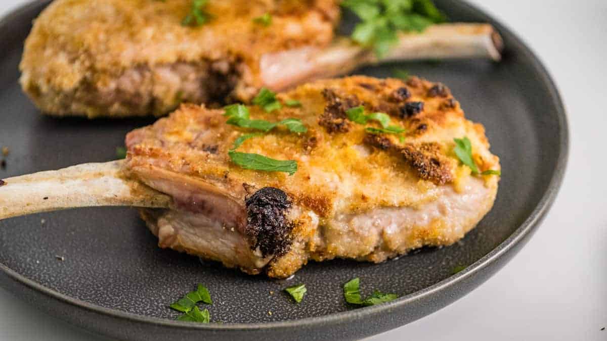 A breaded and cooked pork chop with a bone is served on a dark plate, garnished with chopped parsley—perfect for Everyday Cooking and Quick and Easy dinners.