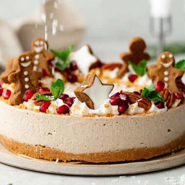A creative cheesecake topped with Gingerbread treats, whipped cream, pomegranate seeds, mint leaves, and star-shaped cookies on a light plate.