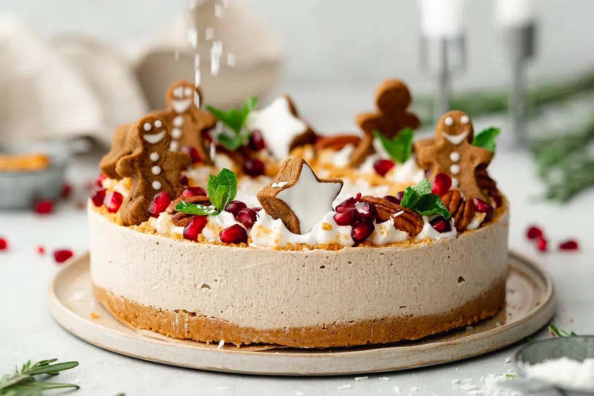 A creative cheesecake topped with Gingerbread treats, whipped cream, pomegranate seeds, mint leaves, and star-shaped cookies on a light plate.