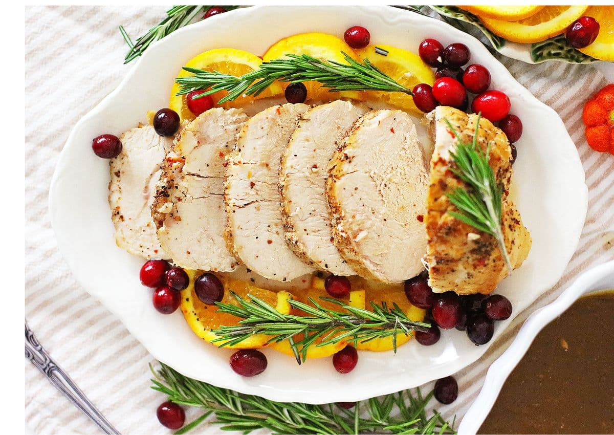 Sliced roasted turkey breast on a white platter, garnished with orange slices, cranberries, and sprigs of rosemary&mdash;perfect for pairing with your favorite slow cooker recipes.