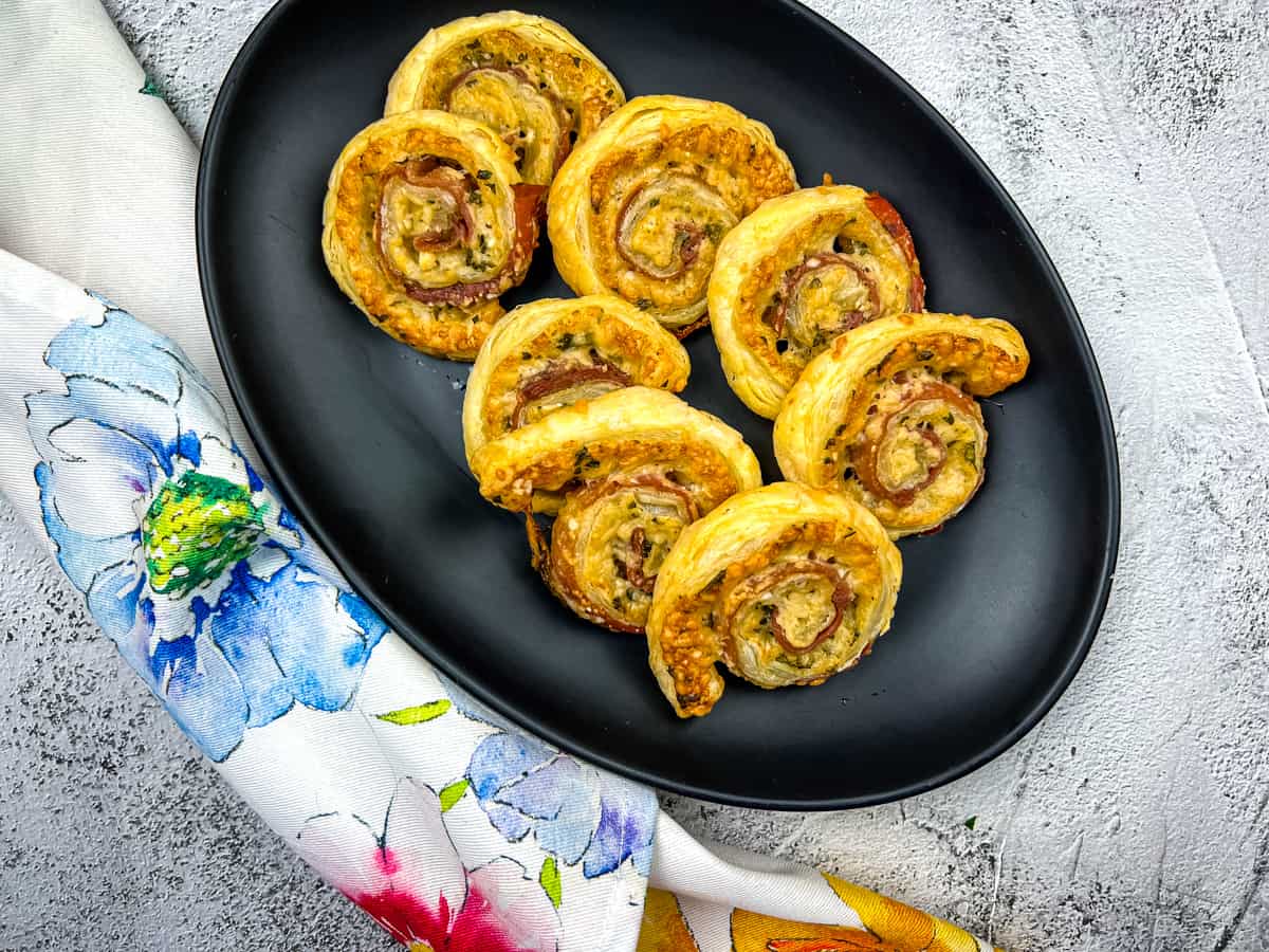 A black oval plate holds eight puff pastry pinwheels filled with ham and cheese, a delicious option for everyday cooking, placed on a textured gray surface with a floral-patterned cloth beside it.