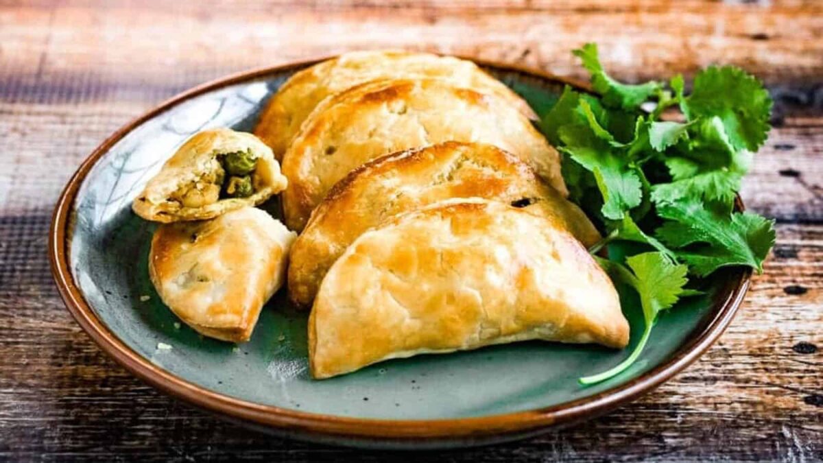 A plate with five baked empanadas, one cut open to show a vegetable filling, sits next to fresh cilantro on a rustic wooden table&mdash;perfect to elevate everyday cooking with easy puff recipes.