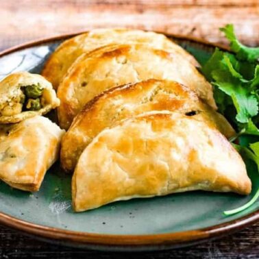 A plate with five baked empanadas, one cut open to show a vegetable filling, sits next to fresh cilantro on a rustic wooden table—perfect to elevate everyday cooking with easy puff recipes.
