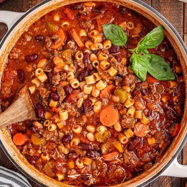 A comforting pot of pasta e fagioli soup with ditalini pasta, beans, ground meat, carrots, and tomatoes—naturally dairy-free and perfect for mains. Fresh basil tops it off, while a wooden spoon rests in the pot.