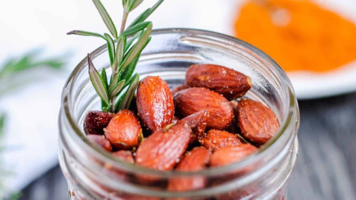 A glass jar filled with roasted almonds garnished with a sprig of rosemary, ideal for joyful vegan dining, with a blurred dish in the background.