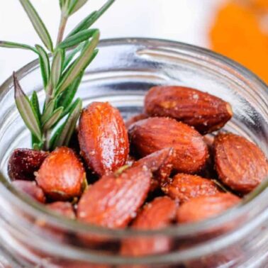 A glass jar filled with roasted almonds garnished with a sprig of rosemary, ideal for joyful vegan dining, with a blurred dish in the background.