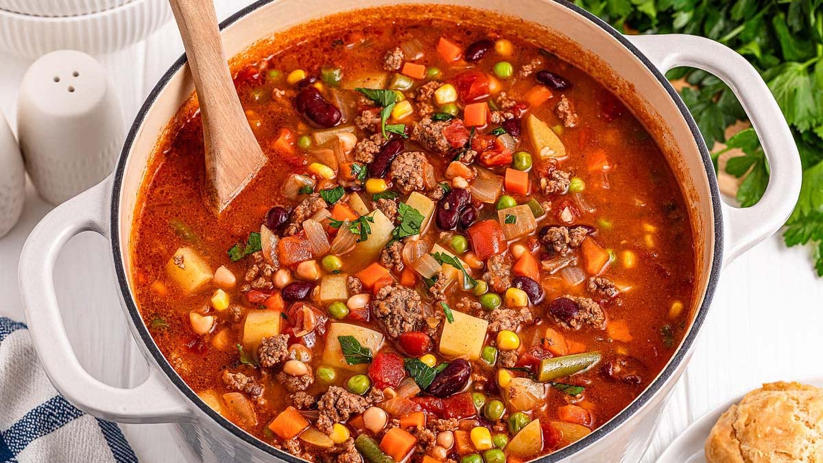 A pot of vegetable and beef soup with beans, peas, corn, potatoes, carrots, and tomatoes is stirred with a wooden spoon on a white surface—one of our favorite comforting mains from classic dairy-free soups.
