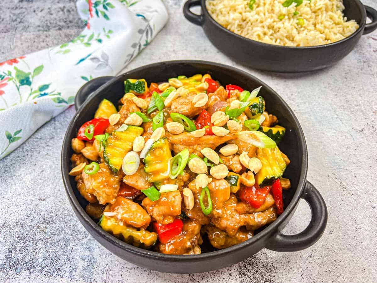 A black bowl filled with stir-fried chicken, vegetables, peanuts, and green onions sits next to rice&mdash;a perfect choice for those seeking flavorful egg-free recipes.