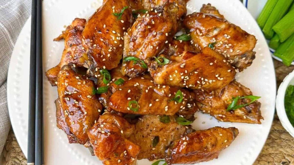 A plate of glazed chicken wings garnished with sesame seeds and chopped green onions, served with a side of celery sticks—perfect for those seeking delicious new chicken wing recipes.
