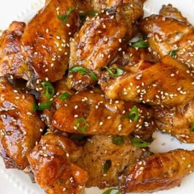 A plate of glazed chicken wings garnished with sesame seeds and chopped green onions, served with a side of celery sticksโperfect for those seeking delicious new chicken wing recipes.