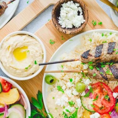 A plate with flavorful gluten-free grilled meat skewers, rice, sliced tomatoes, olives, feta cheese, hummus, and a side of salad is shown on a table.