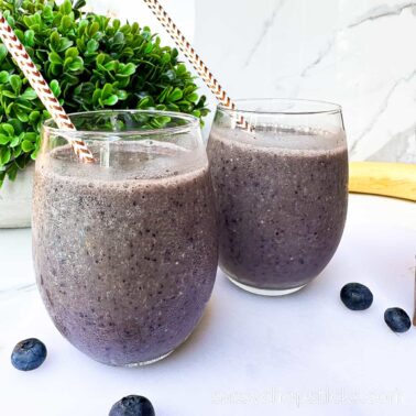Two glasses of blueberry smoothie with straws on a white surface, garnished with a plant and scattered blueberries—one of the best drinks to brighten any moment.