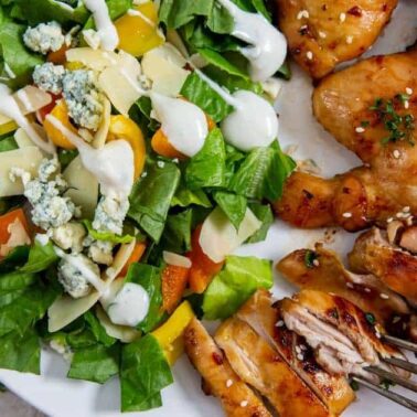 A plate with juicy chicken thigh, a green salad with cheese, vegetables, and dressing, and a fork holding a piece of tender chicken.