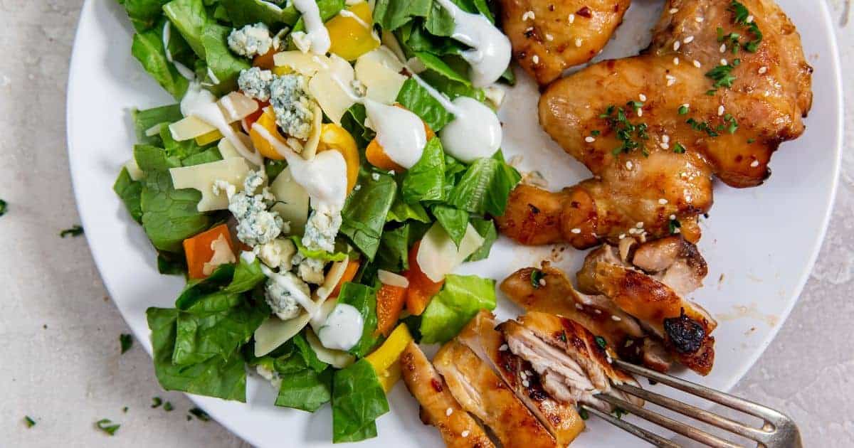 A plate with juicy chicken thigh, a green salad with cheese, vegetables, and dressing, and a fork holding a piece of tender chicken.