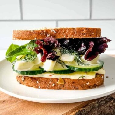 A vegetable sandwich with leafy greens, cucumber, cheese, and herbs on whole grain bread, served on a white plate—perfect for anyone seeking new sandwich ideas or the best sandwiches to enjoy.