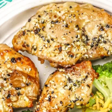 Three pieces of sesame-crusted, juicy chicken thighs on a white plate with a side of steamed broccoli and shredded cheeseโperfect for those seeking bold flavor in their chicken thigh recipes.