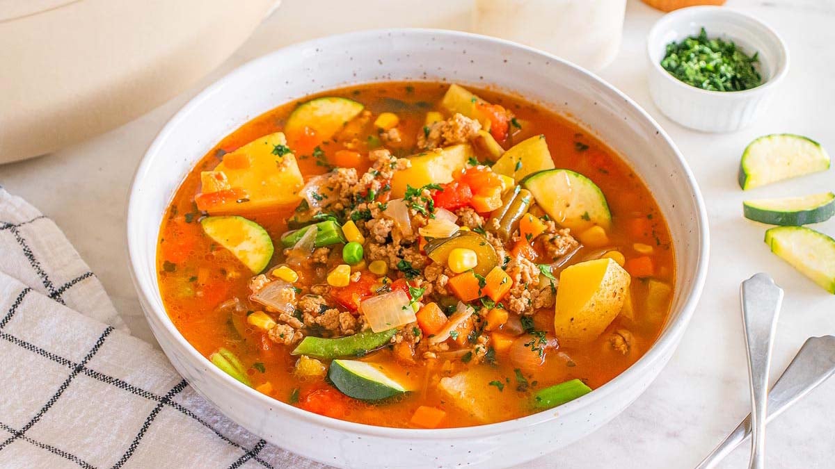 A bowl of vegetable soup with ground meat, potatoes, carrots, corn, peas, green beans, and zucchini in a tomato broth sits on a marble surface beside a napkin and utensils—perfect for simple eating and delicious everyday eating.
