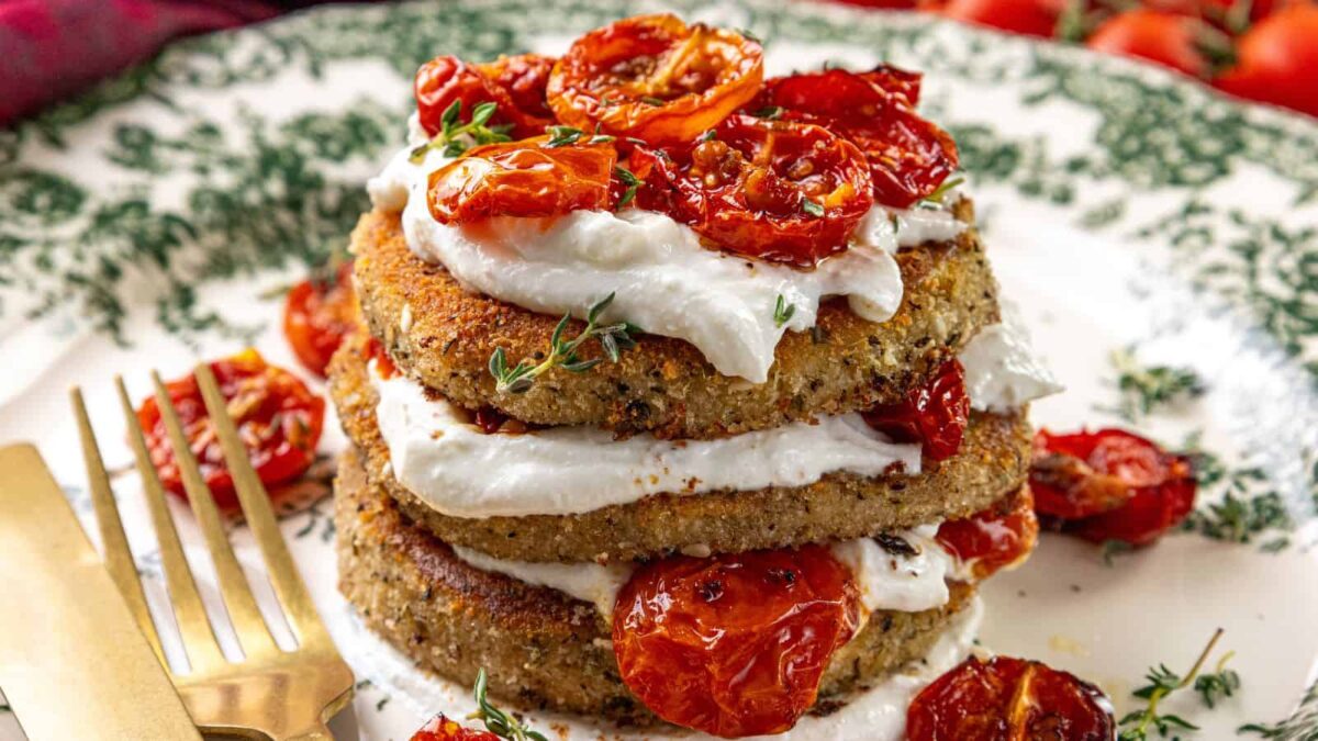 Three stacked breaded patties layered with creamy cheese and roasted tomatoes, garnished with herbs&mdash;an elegant addition to ricotta appetizers&mdash;served on a decorative plate with a fork and knife.