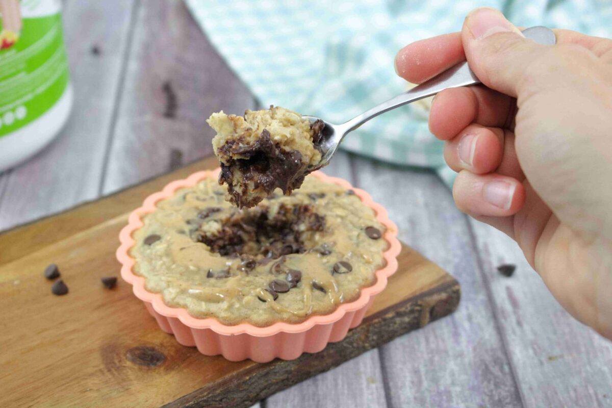A hand holds a spoonful of chocolate chip mug cake above a pink silicone ramekin filled with the dessert, on a wooden board&mdash;perfect for quick recipes or adding Oats for wholesome meal ideas.