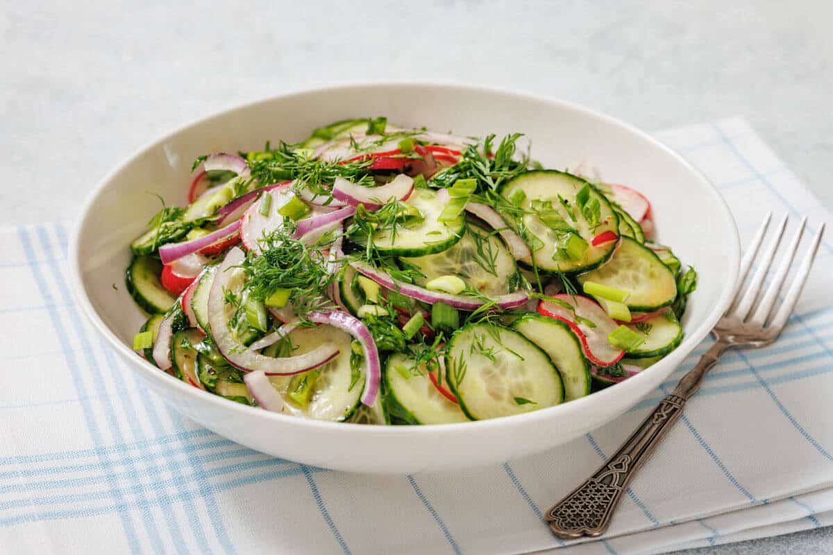 A white bowl filled with cucumber salad containing sliced cucumbers, red onions, radishes, and fresh herbs sits on a white and blue cloth with a fork beside it—a perfect choice for simple recipes and everyday eating.