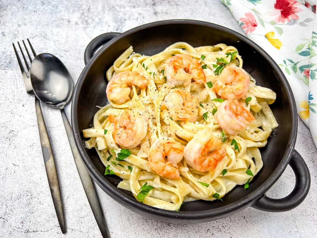 A black bowl filled with creamy fettuccine pasta topped with shrimp and chopped parsley, served beside a fork and spoon on a light surface&mdash;perfect for recreating restaurant favorites at home.