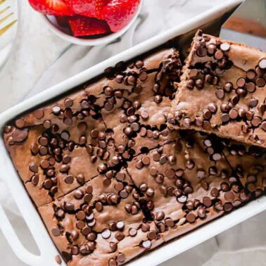 A rectangular dish filled with chocolate chip brownies made with oats, some pieces cut and stacked, with a bowl of sliced strawberries nearbyโperfect for new recipes or sweet meal ideas.