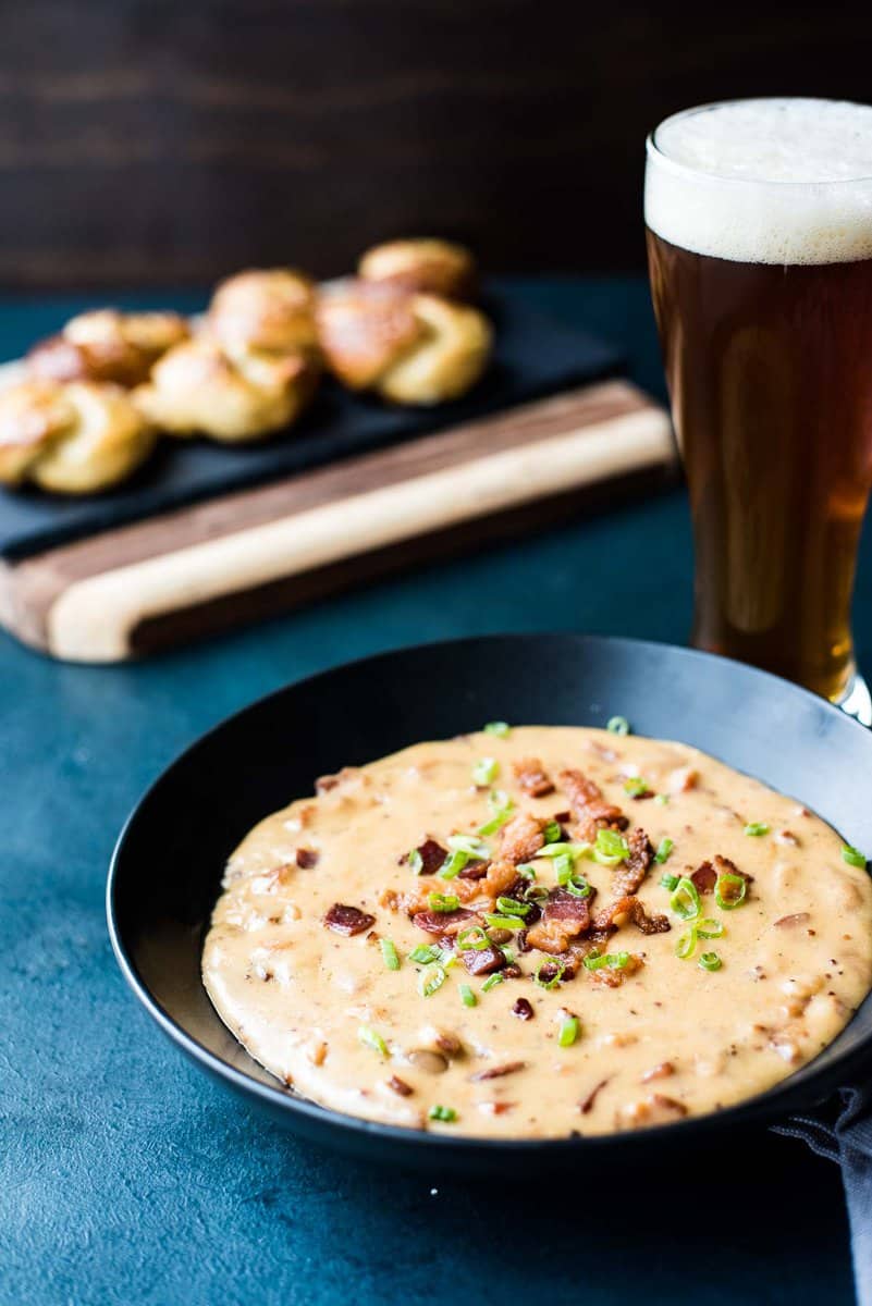 A black bowl of creamy soup topped with chopped bacon and green onions, served next to a glass of beer and bread rolls&mdash;perfect for those seeking delicious Bacon Appetizer Ideas or new Bacon Recipes.