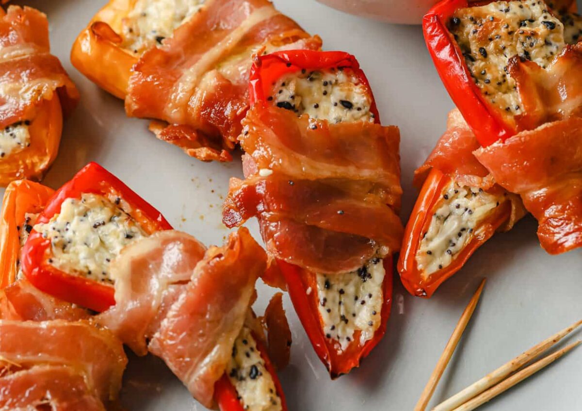 Mini red peppers stuffed with a creamy cheese mixture and wrapped in bacon are arranged on a plate next to toothpicks&mdash;perfect for those searching for delicious bacon appetizers.