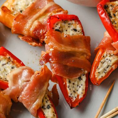 Mini red peppers stuffed with a creamy cheese mixture and wrapped in bacon are arranged on a plate next to toothpicks—perfect for those searching for delicious bacon appetizers.