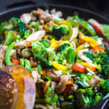 A close-up of a skillet filled with stir-fried chicken, broccoli, bell peppers, and other mixed vegetables showcases one of the most satisfying dinnersโperfect for fans of easy broccoli recipes. A wooden spatula rests on the side.