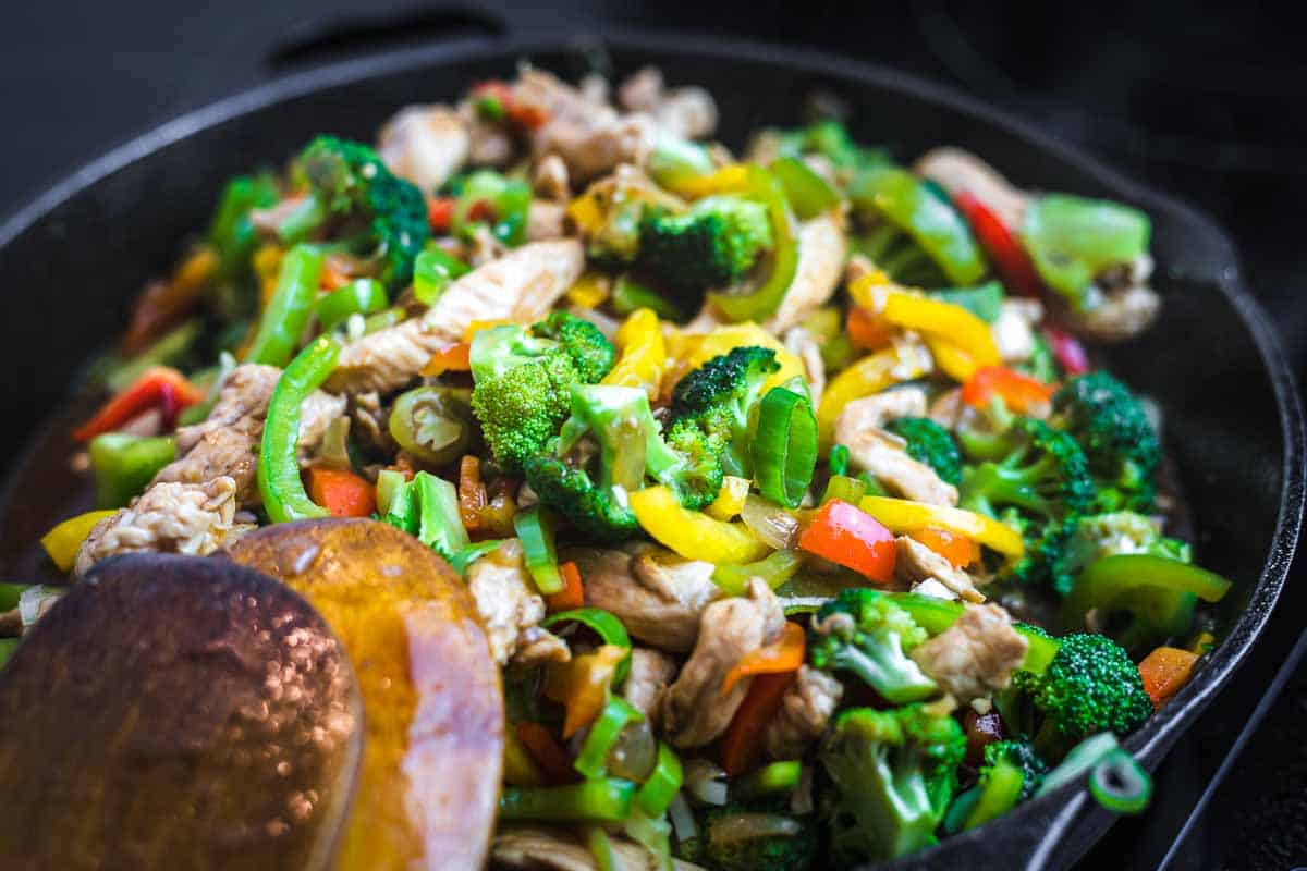 A close-up of a skillet filled with stir-fried chicken, broccoli, bell peppers, and other mixed vegetables showcases one of the most satisfying dinners&mdash;perfect for fans of easy broccoli recipes. A wooden spatula rests on the side.