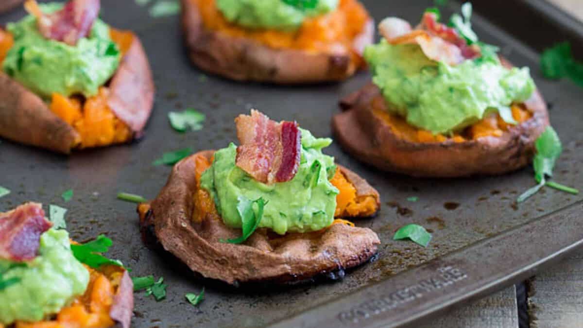 Baked sweet potato rounds topped with guacamole, crispy bacon pieces, and chopped herbs on a baking sheet&mdash;a tasty twist on classic bacon recipes.