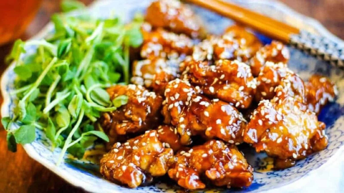 A plate of glazed sesame chicken pieces garnished with white sesame seeds, served with fresh green herbs and chopsticks&mdash;a perfect choice for quick Chicken Dinner Ideas or a Fast Dinner using your Air Fryer Chicken recipe.