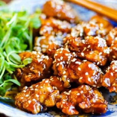 A plate of glazed sesame chicken pieces garnished with white sesame seeds, served with fresh green herbs and chopsticks—a perfect choice for quick Chicken Dinner Ideas or a Fast Dinner using your Air Fryer Chicken recipe.