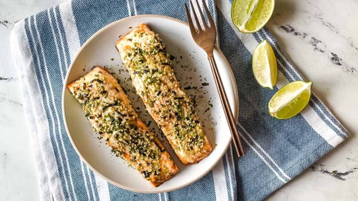 Two baked salmon fillets topped with herbs and spices on a white plate, with a fork, knife, and lime wedges on a striped kitchen towel&mdash;perfect for an umami-rich Asian-style dinner.