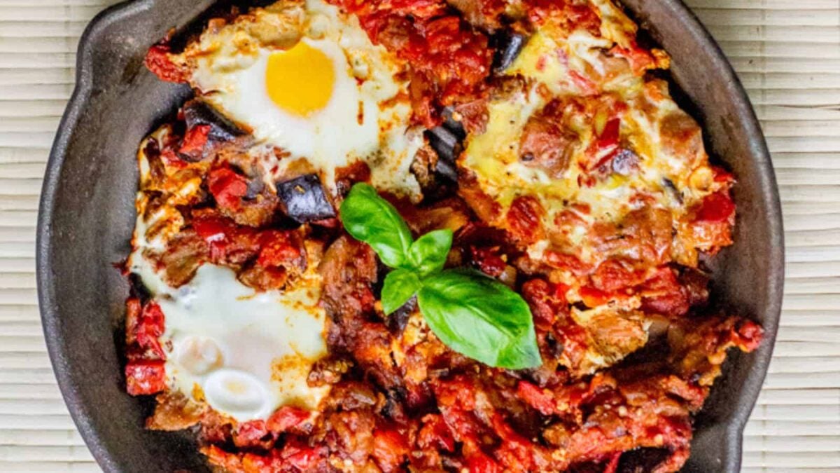 A skillet filled with shakshuka: poached eggs in a tomato and vegetable sauce, garnished with a sprig of fresh basil&mdash;perfect for hearty vegetarian breakfast ideas.