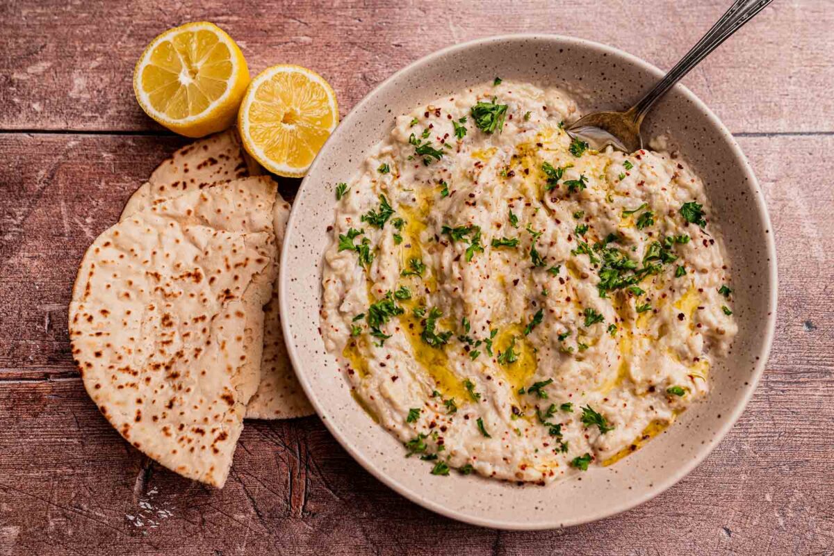 A bowl of baba ganoush&mdash;a classic among delicious eggplant recipes&mdash;garnished with parsley and olive oil, accompanied by lemon halves and flatbread on a wooden surface.