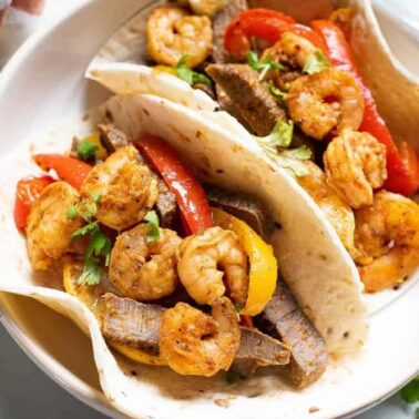 Two flour tortillas filled with shrimp, beef strips, and bell peppers sizzle with flavor, garnished with cilantro for a touch of comfort—an irresistible take on classic fajita recipes served on a round white plate.