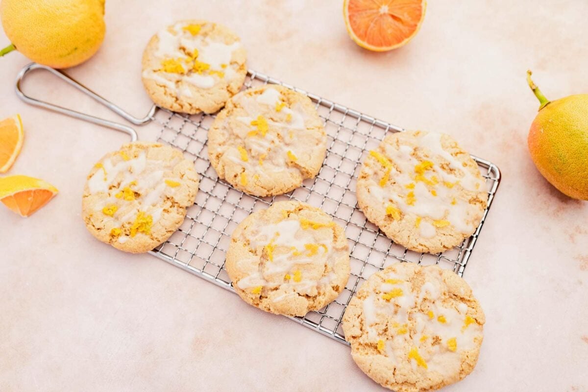 Six glazed citrus cookies, a delightful gluten-free dessert, are arranged on a cooling rack and surrounded by fresh citrus fruits and slices on a light-colored surface.