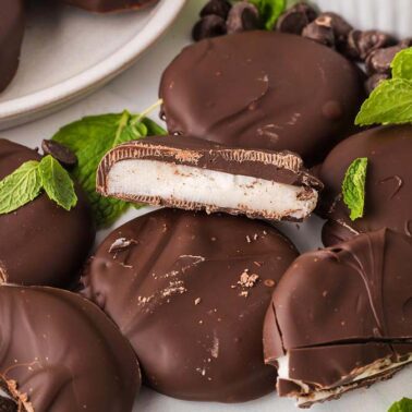 Chocolate-covered peppermint patties are arranged on a white surface, some cut open to reveal a white mint filling. Fresh mint leaves surround these sweet creations, making them a delightful option for lovers of gluten-free desserts.