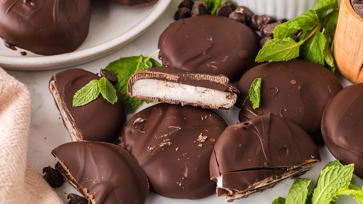 Chocolate-covered peppermint patties are arranged on a white surface, some cut open to reveal a white mint filling. Fresh mint leaves surround these sweet creations, making them a delightful option for lovers of gluten-free desserts.