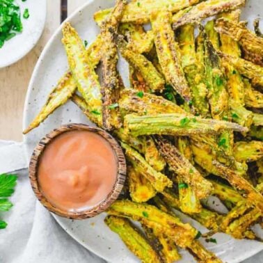 A plate of crispy baked okra fries, perfect as a weeknight side dish, garnished with herbs and served with a small wooden bowl of dipping sauce. Fresh parsley and chopped herbs are nearby.
