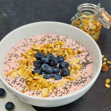 A bowl of creamy yogurt tinted pink, topped with blueberries, chia seeds, and sliced almonds, sits beside a small jar of almonds and loose blueberries on a dark surface—a delicious treat perfect for versatile yogurt recipes.