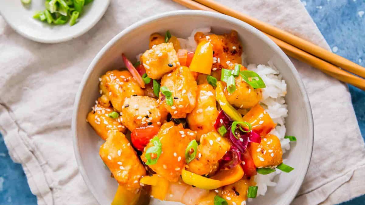 A bowl of white rice topped with sweet and sour chicken, colorful bell peppers, chopped green onions, and sesame seeds—this pantry staple makes real meals simple. Chopsticks and a small dish of green onions sit beside the bowl.