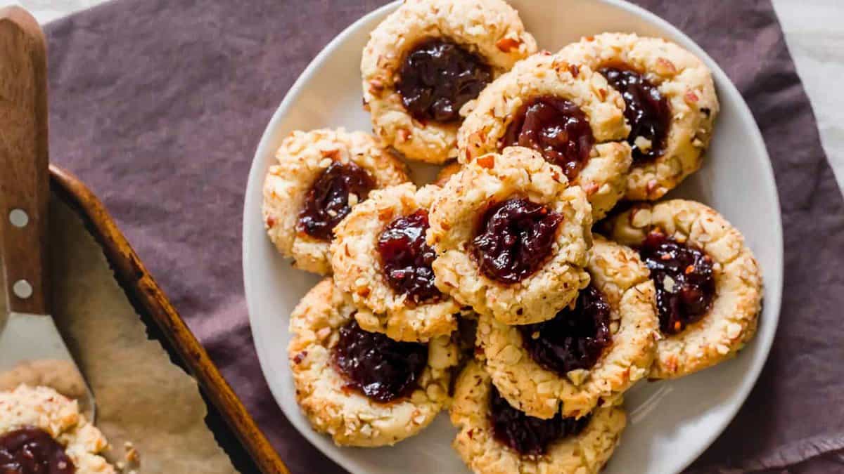 A plate of freshly baked Thumbprint cookies filled with red jam sits on a white plate, with a baking tray partially visible beside it—perfect for trying out new cookie recipes.