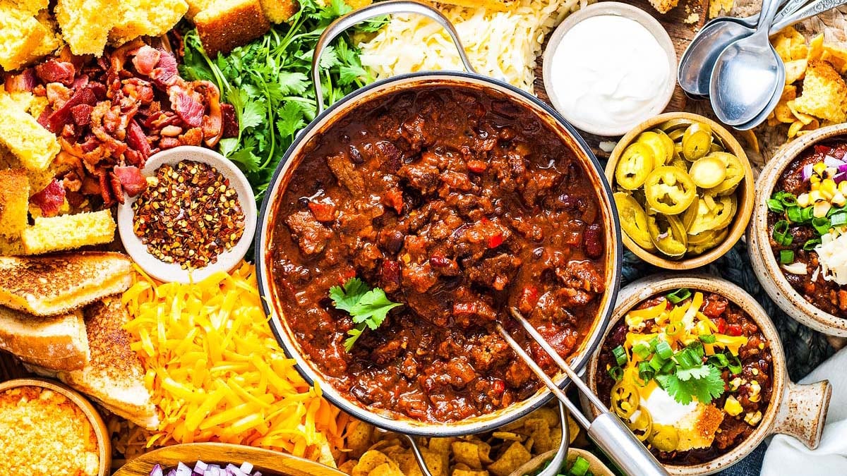 A bowl of chili surrounded by various toppings like shredded cheese, jalapenos, cornbread, bacon, sour cream, cilantro, and chopped onions—perfect for weeknight meals or anyone seeking comforting recipes.
