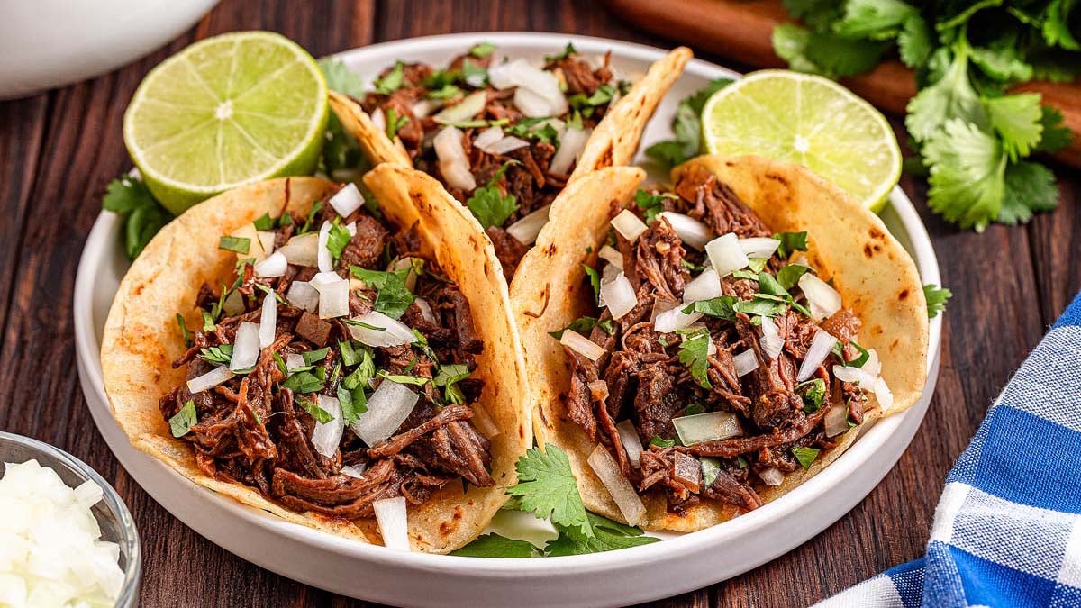 Three shredded beef tacos topped with chopped onions and cilantro, served on a white plate with lime halves and fresh cilantro in the background—a perfect choice for flavorful dinner or gluten-free recipes.