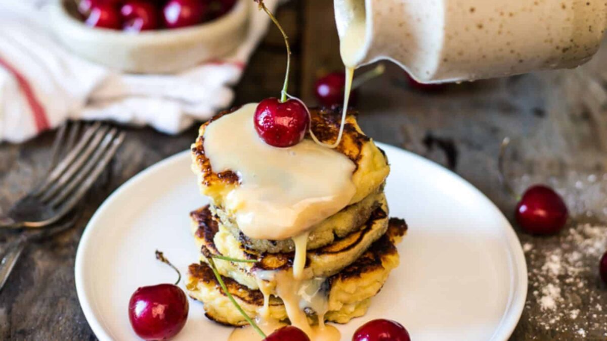 A stack of pancakes topped with cherries is being drizzled with a creamy sauce, served on a white plate with extra cherries&mdash;a perfect treat for feel-good mornings and lovers of fluffy pancake recipes.