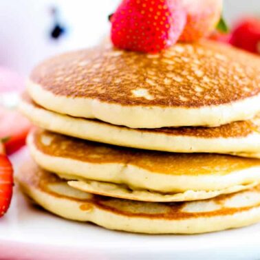 A stack of three pancakes on a white plate, topped with whole and sliced strawberries—perfect for feel-good mornings and anyone who loves fluffy pancake recipes.